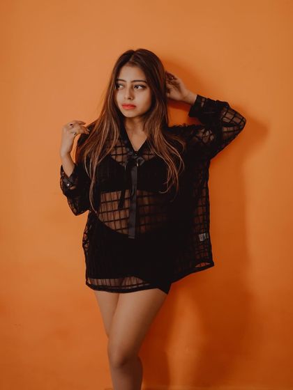 A bold studio portrait using a bright orange background to create a high-impact fashion image. The sheer black top adds a layer of texture and style.