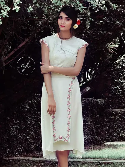 An elegant white indie-chic dress featuring delicate pink embroidery along the side and on the cap sleeves. The asymmetrical hemline and pleated bodice details create a modern silhouette rooted in traditional aesthetics.