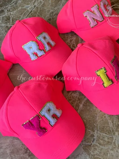 Caps with a personal touch. These vibrant neon pink baseball caps are perfect for a bachelorette party or group event, each customized with two-letter initials in sparkling patches.