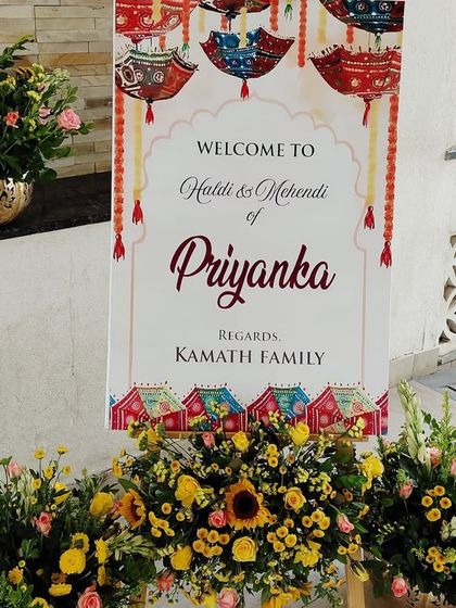 This is a personalized welcome sign we designed for a Haldi and Mehendi function. The sign features thematic graphics and is surrounded by arrangements of yellow flowers and sunflowers to match the event's decor.