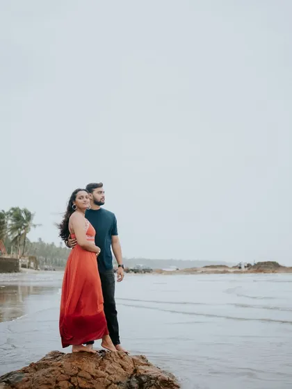 A couple stands on a rock by the sea, looking out at the horizon. A beautiful, contemplative shot from a beach pre-wedding photoshoot.