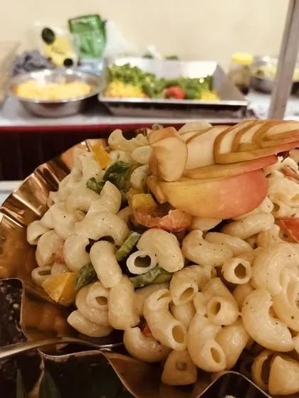 A creamy macaroni salad with mixed vegetables, garnished with fanned apple slices and served in a decorative golden bowl.
