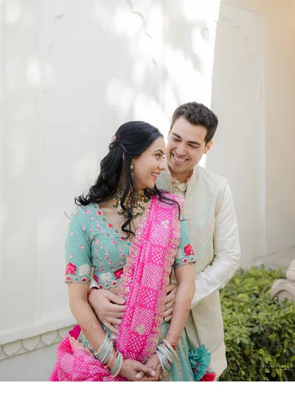 Sharing a happy, candid moment. The vibrant colors of their traditional outfits pop against the clean white walls of the Jaipur hotel, creating a fresh and joyful pre-wedding portrait.