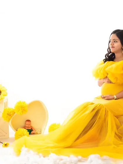 A bright and cheerful studio setup with a mom-to-be in a sunny yellow gown. The scene is decorated with sunflowers and white clouds, creating a whimsical and airy high-key maternity portrait.