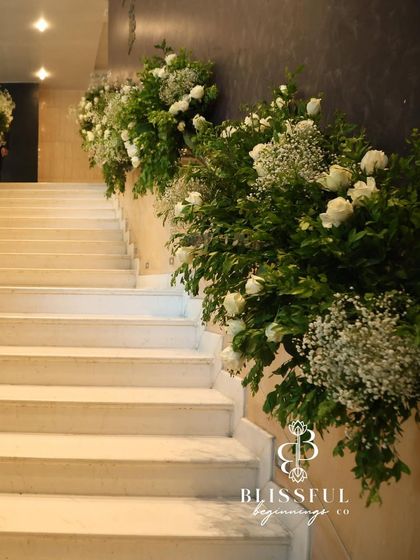 A close-up of the staircase decor, showing the lush combination of white roses, baby's breath, and various types of greenery. The arrangement feels organic and abundant, yet incredibly elegant.