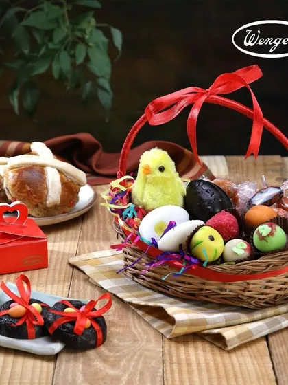An Easter basket filled with joy. This wicker basket is brimming with our handmade marzipan eggs, assorted chocolates, a little toy chick, and our freshly baked hot cross buns in the background.