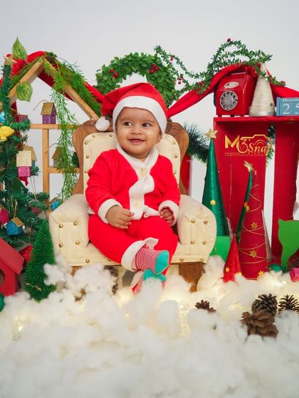 A festive Christmas-themed baby shoot. This little one looks adorable dressed as Santa, sitting in a miniature armchair surrounded by snow and holiday decorations.