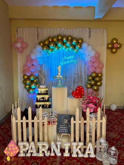 A simple and elegant setup with a white curtain backdrop and a gold and pink balloon arch. The decor is accented with a neon sign, a picket fence, and marquee letters.