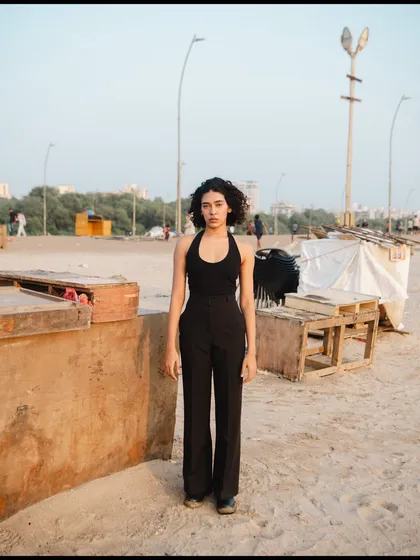 A full-length portrait with a raw, almost documentary feel. The industrial beach background contrasts with the model's simple, chic outfit.