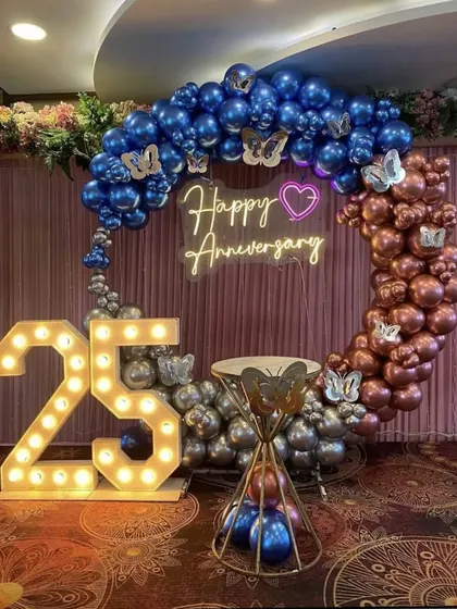 This vibrant 25th anniversary decoration uses a striking color combination of chrome blue and rose gold. The setup is complete with a circular balloon frame, large marquee numbers, and a "Happy Anniversary" neon sign for a modern party look.