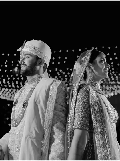 A classic black and white portrait of the couple standing back-to-back. Their regal attire and confident poses are beautifully highlighted against the sparkling lights.