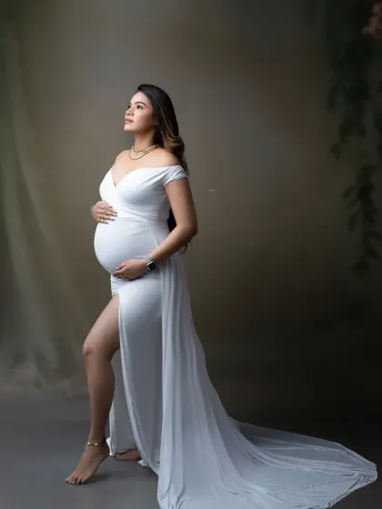 A classic and elegant maternity pose. The simple white gown with a long train accentuates the beauty of the baby bump against a soft, neutral background.