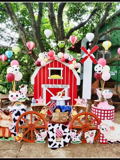 I design charming and unique themes like this farmyard party. The setup features a classic red barn backdrop, adorable cutouts of farm animals like cows and sheep, wagon wheel props, and even hot air balloon decorations hanging from the trees for a touch of whimsy.