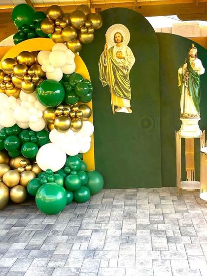 A First Holy Communion decoration in green, white, and gold. The setup includes backdrops with religious figures and a matching balloon arrangement.