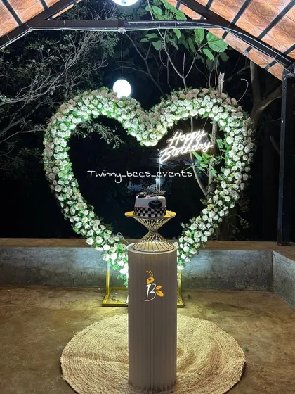 An elegant and romantic birthday or anniversary setup. This features a heart-shaped arch made of white flowers with a "Happy Birthday" neon sign, perfect for a surprise celebration at night.