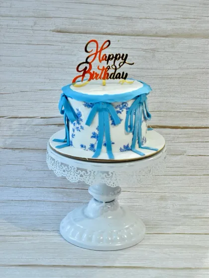 An elegant birthday cake with a vintage feel. This design features delicate blue floral patterns and charming fondant ribbon bows on a smooth white surface.
