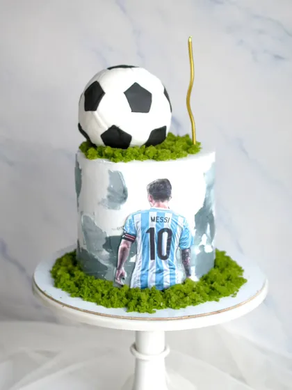A football cake for a Messi fan. This design features an edible print of the player, a 3D soccer ball on top, and green cake-crumb "grass" for a realistic stadium feel.