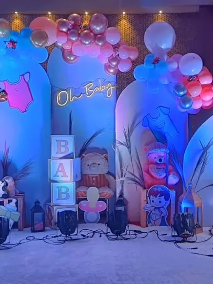 A baby shower setup with blue arched backdrops, a neon "Oh Baby" sign, and props like "BABY" blocks and a teddy bear.