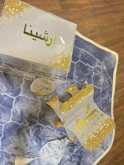 A personalized gift set featuring a dome-shaped prayer mat in a beautiful blue pattern. The set is completed with a custom resin Quran box and holder, a crystal tasbeeh, a bookmark, and a car hanging, all in a coordinated white and gold glitter design.