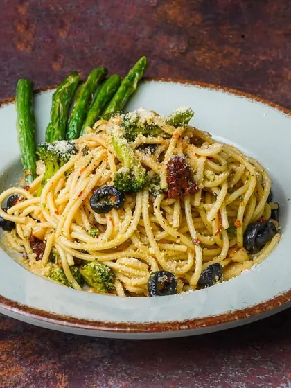 A plate of aglio e olio spaghetti with broccoli and black olives, shot on a dark, textured surface. This styling gives a classic dish a sophisticated and rustic feel.