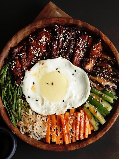 A delicious and hearty meal in a bowl, featuring Korean-style grilled meat, a sunny-side-up egg, and an assortment of fresh vegetables over rice.