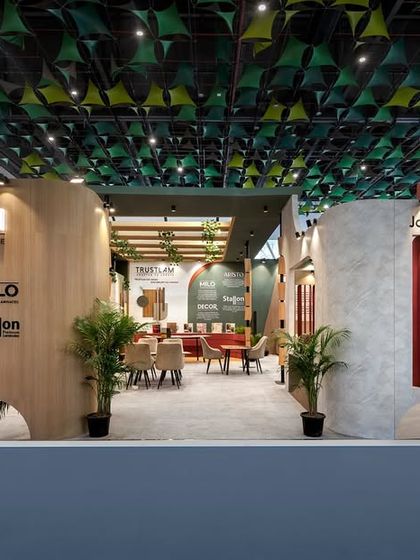 The entrance to the Trustlam pavilion is designed to be open and accessible, with clear branding and a glimpse of the inviting interior. The design uses a mix of wood, concrete textures, and greenery to reflect a modern yet earthy brand identity.