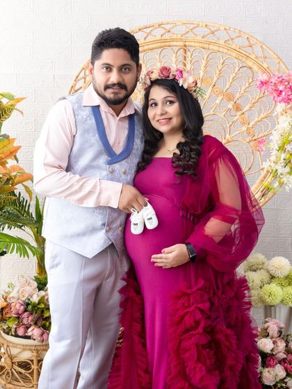 A proud portrait of the parents-to-be. Dressed in vibrant magenta, they hold up a pair of baby shoes, their smiles full of excitement for the new arrival.