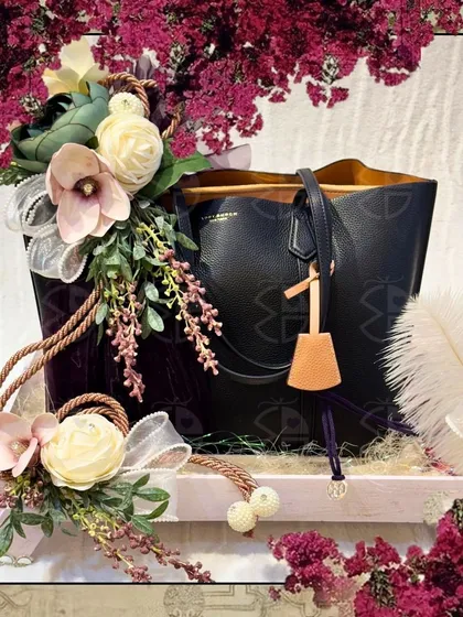 A classic black tote bag, elegantly presented on a rustic wooden tray. The arrangement is enhanced with delicate floral bouquets and a feather, adding a touch of softness to the design.