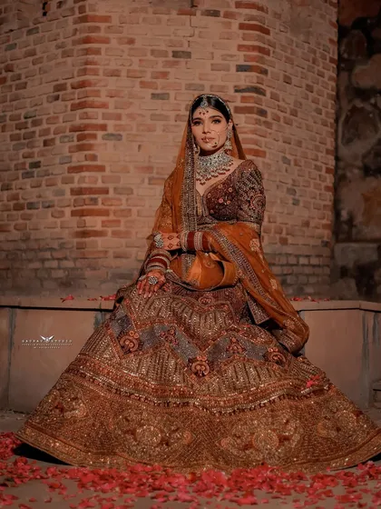 A full-shot of the bride seated against a rustic brick wall, her ornate brown lehenga spread beautifully around her.