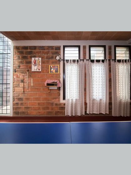 The blue of a table tennis table adds a splash of color to this family room, which is otherwise defined by the earthy tones of its brick walls and red flooring. Simple white curtains diffuse the light from the windows.