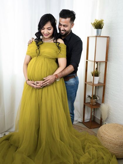 A happy and relaxed couple's portrait in our studio. The partner embraces the mom-to-be from behind, both smiling and looking down at her beautiful baby bump.