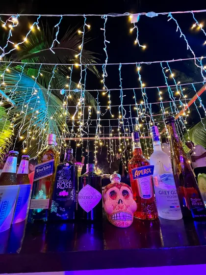 A fully stocked bar at night, illuminated by hanging fairy lights. The centerpiece is a decorative skull bottle, a nod to our edgy brand identity, surrounded by a premium selection of spirits.