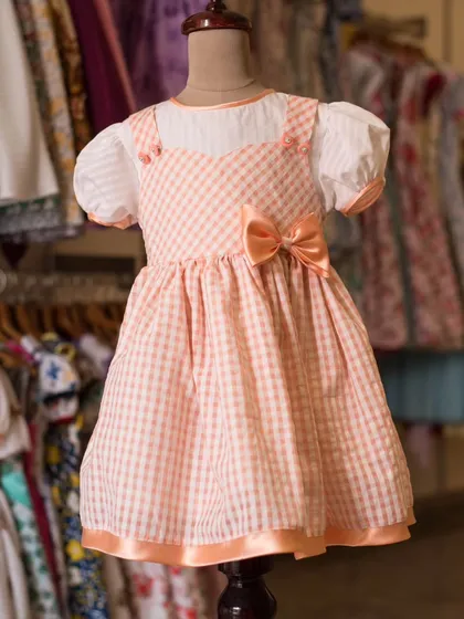 This sweet peach-colored gingham pinafore dress is layered over a blouse with puff sleeves. It's a classic, charming look that is perfect for any casual outing.