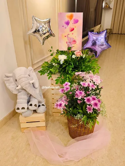 A sweet corner setup for a baby girl's arrival. It features a plush elephant toy, star-shaped balloons, and baskets of fresh pink and white flowers, creating a soft and celebratory welcome.