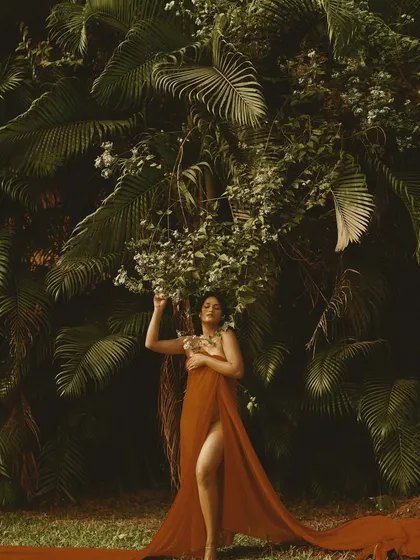 A wide shot that shows the full effect of the flowing red fabric against the lush green background. The hair and makeup are designed to feel organic and part of the natural world.