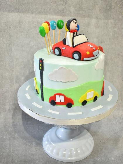 A full view of the car-themed cake on a stand, showing the road wrapping around the base and the colorful balloon toppers.