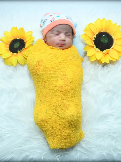 A little ray of sunshine. This newborn is wrapped in a cheerful yellow swaddle, sleeping peacefully between two large sunflower props on a soft white fur rug.
