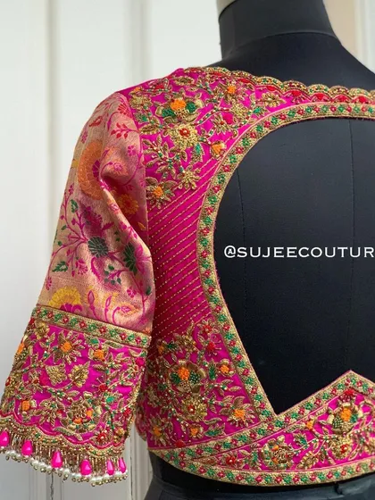 A stunning pink bridal blouse with a unique back design. The sleeves are made from a floral brocade fabric, while the back features a large cutout framed by multi-colored zardozi and beadwork.