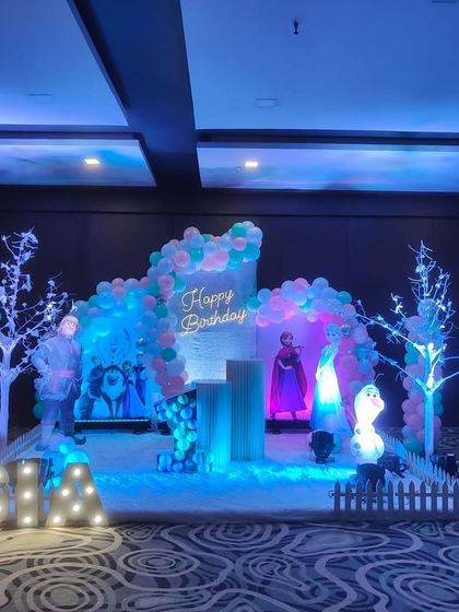 A "Frozen" themed winter wonderland for Sia's first birthday. The stage is decorated to look like a snowy landscape with light-up trees, character cutouts of Anna and Elsa, and cool blue lighting.