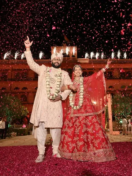 The couple raises their hands in celebration after a successful varmala exchange, with a shower of petals raining down on them in front of a grand palace.