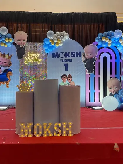 A large-scale 'Boss Baby' themed first birthday party on a stage. This setup includes multiple character cutouts, custom backdrops, marquee letters spelling the baby's name, and extensive balloon work in shades of blue and silver.