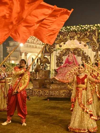 The bride makes her grand entrance on a beautiful palanquin, framed by dancers waving large orange flags. This wide shot captures the impressive scale and traditional grandeur of the moment.
