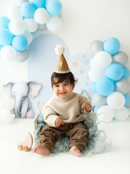 A smiling baby boy in a cozy sweater and party hat, celebrating his first birthday in the studio.
