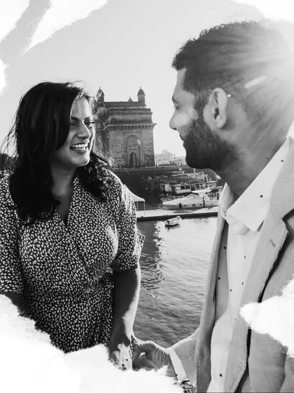 A bright, high-contrast black and white photo of a couple enjoying a moment by the sea, with the Gateway of India in the background.