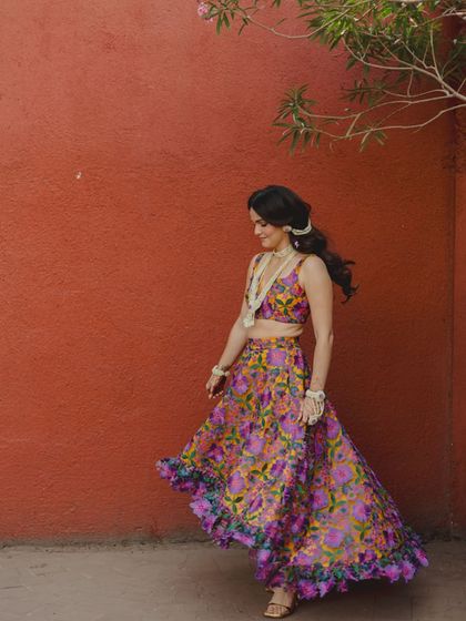 A joyful, twirling portrait of the bride in her vibrant floral lehenga. The red wall provides a perfect, bold backdrop for this happy and energetic Haldi day shot.