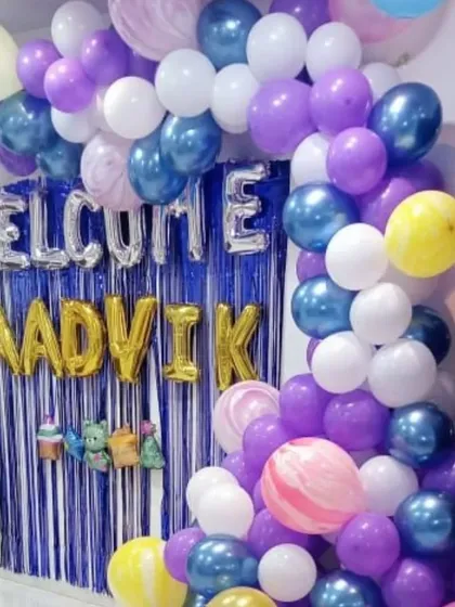 A personalized "Welcome Aadvik" home decoration. This setup includes a blue fringe backdrop, custom name balloons, and a colorful balloon arch in purple, blue, white, and yellow.