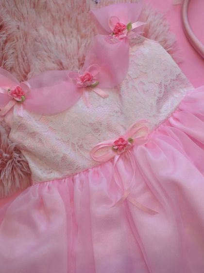 A fairy-tale dream in soft pink. This dress features a delicate lace bodice and layers of sheer fabric, adorned with tiny rosebuds and organza bows on the shoulders for an ethereal, magical look.