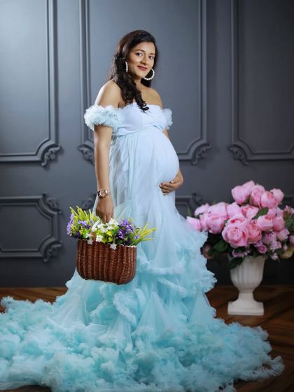 A serene and classic maternity portrait. The mom-to-be is wearing a light blue off-shoulder gown against a classic grey paneled wall in my studio, holding a basket of flowers.