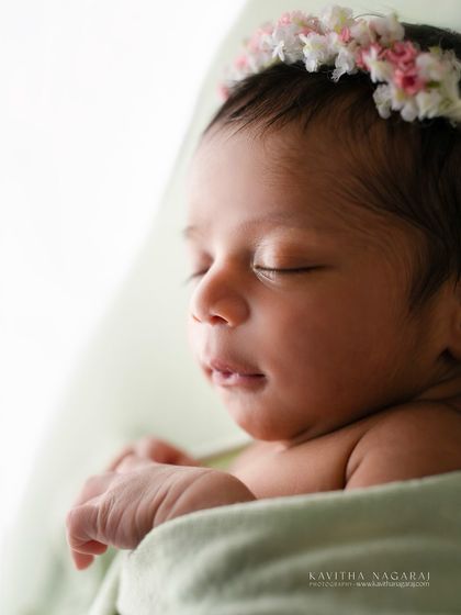 A close-up portrait of a newborn with a delicate floral crown, captured against a soft, bright light. The focus is on the tiny, perfect details of her face.