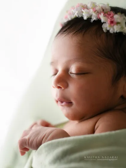 A close-up portrait of a newborn with a delicate floral crown, captured against a soft, bright light. The focus is on the tiny, perfect details of her face.